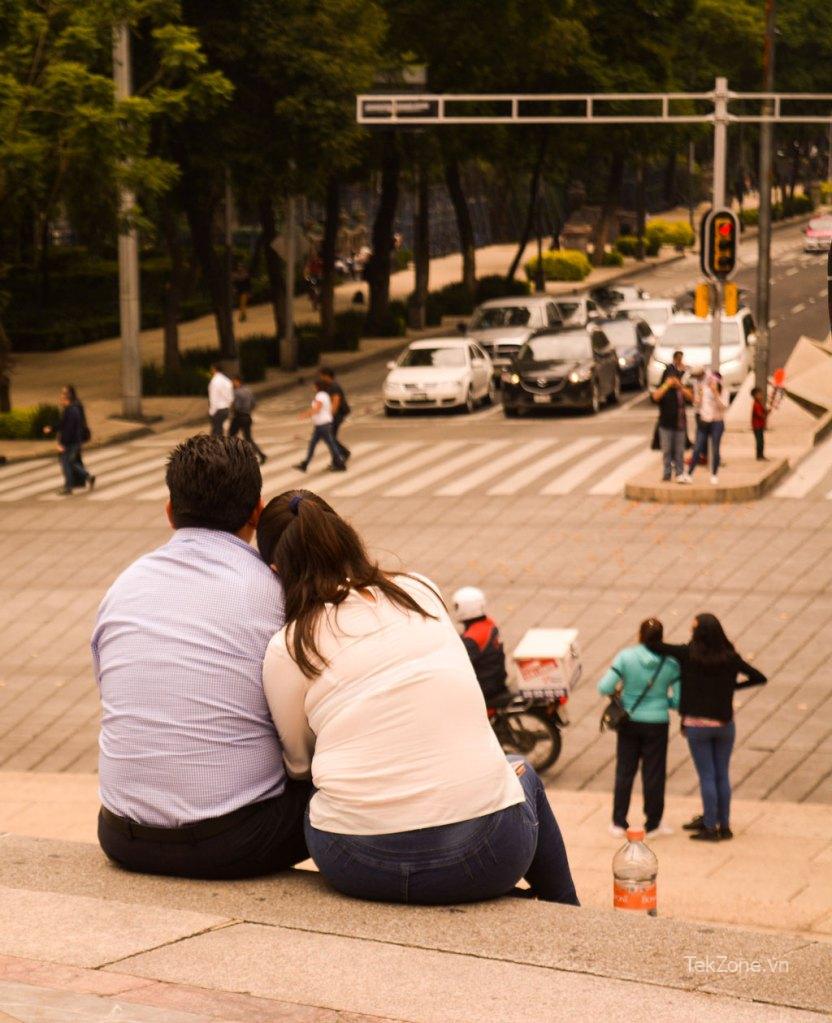 Một cặp đôi ngồi trên bậc thềm của bức tượng El Angel de la Independencia ở Thành phố Mexico, đối diện với một con phố đông đúc, những bức chân dung chân thực