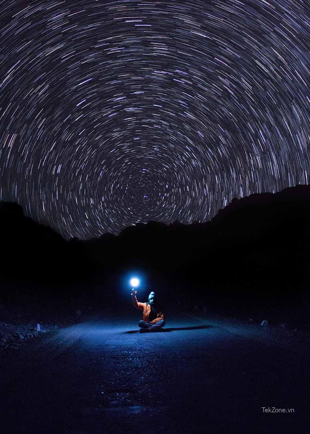 Bầu trời đêm với những vệt sao tròn có hình bóng của một ngọn đồi.  Phía trước có một người ngồi bắt chéo chân trên đường, tay cầm đèn.