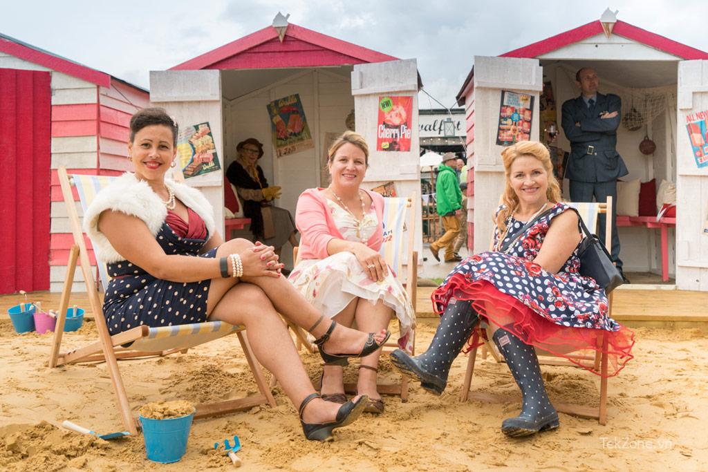 Ba người phụ nữ mặc trang phục cổ điển ngồi trước cabin trên bãi biển.  Ví dụ tô màu Photoshop AI