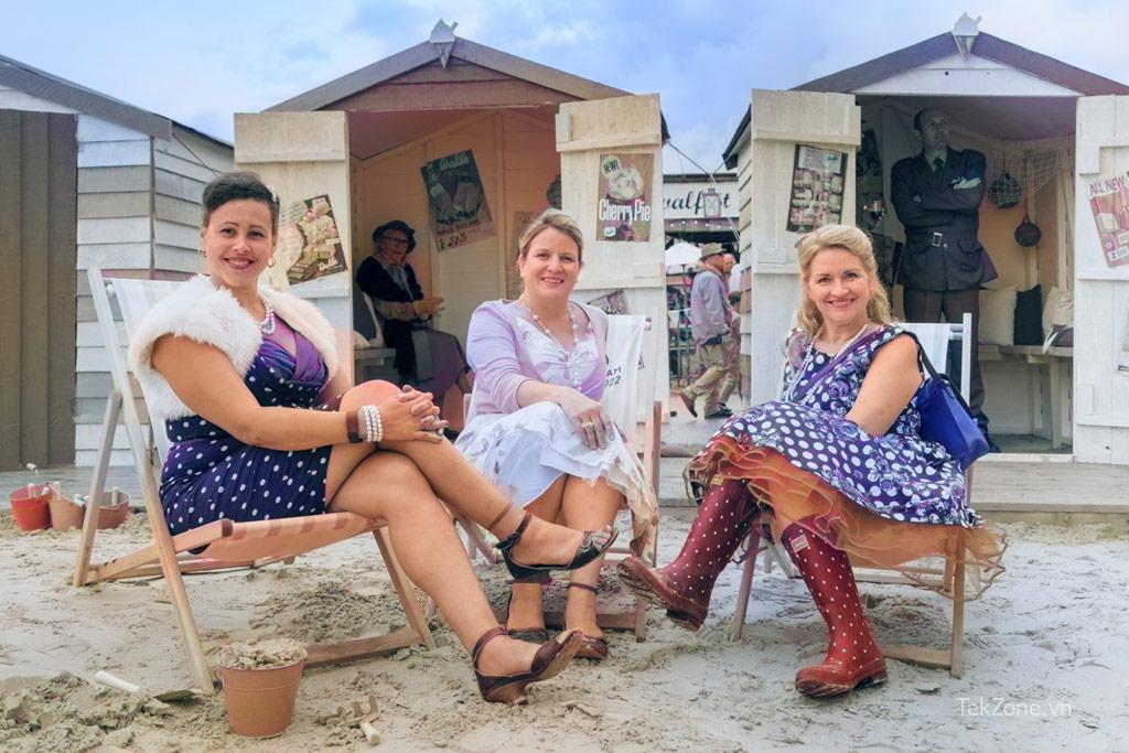 Ba người phụ nữ mặc trang phục cổ điển ngồi trước cabin trên bãi biển.  Ví dụ tô màu Photoshop AI